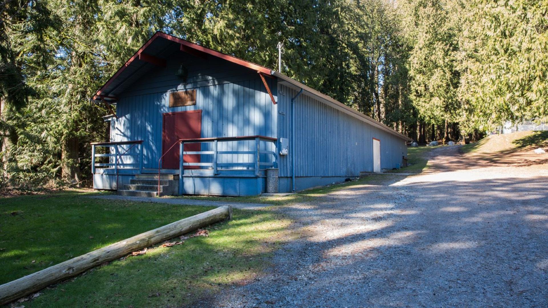 A narrow and long blue, wood sided building. It sits in a wooded area next to a gravel parking lot. 